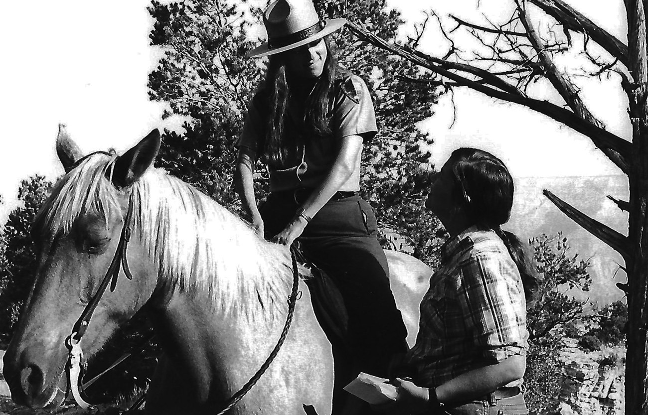 Eileen Szychowski on horseback 2, 1982 Eileen Szychowski in NPS uniforms sits horseback, smiling and looking down at a person standing beside her.
