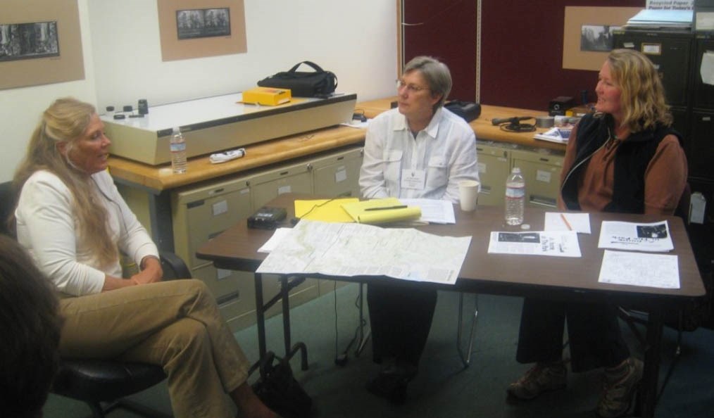 Interviewing NPS employees Two people sit at a table with papers, maps, and a recorder to interview a third person, seated to the left.