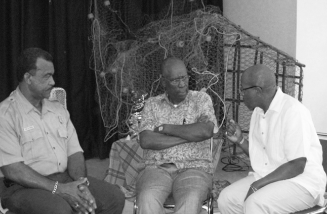 A Black man in NPS uniform listens to two Black men. They are seated in front of fishing nets.