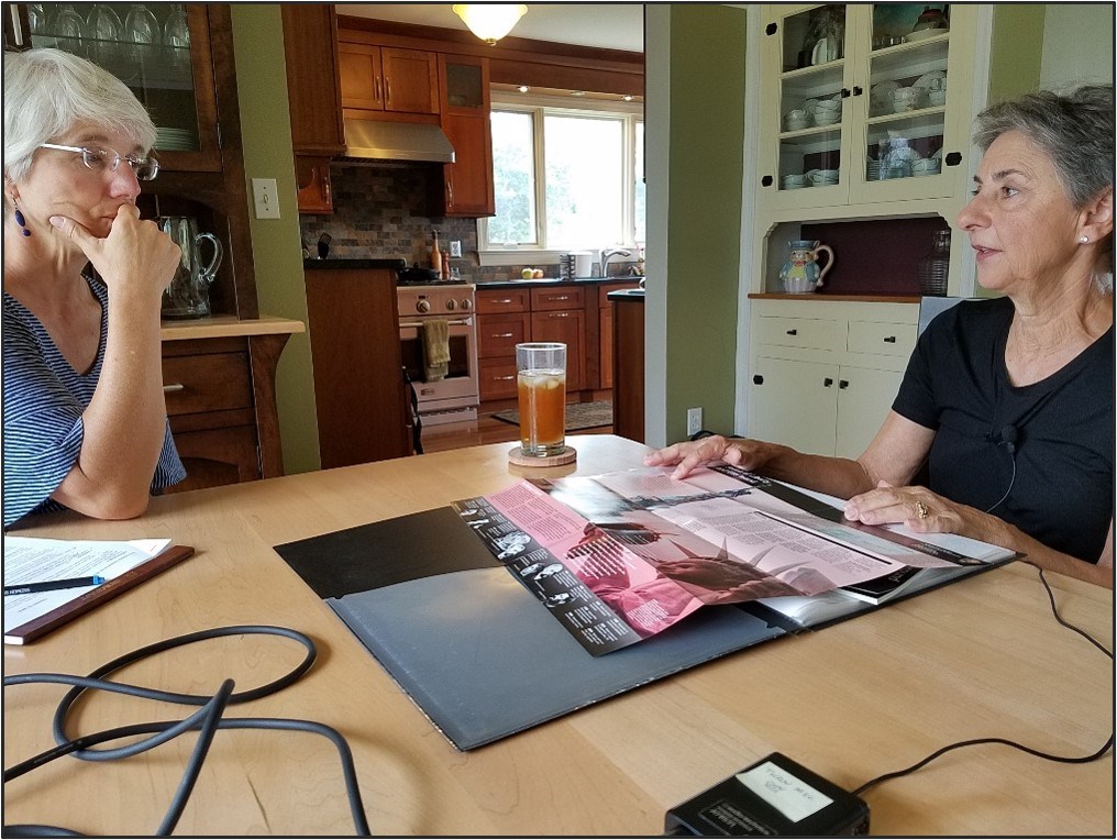 Two people sit at a kitchen table during an oral history interview, with a microphone, beverage glass, and NPS brochure on the table.
