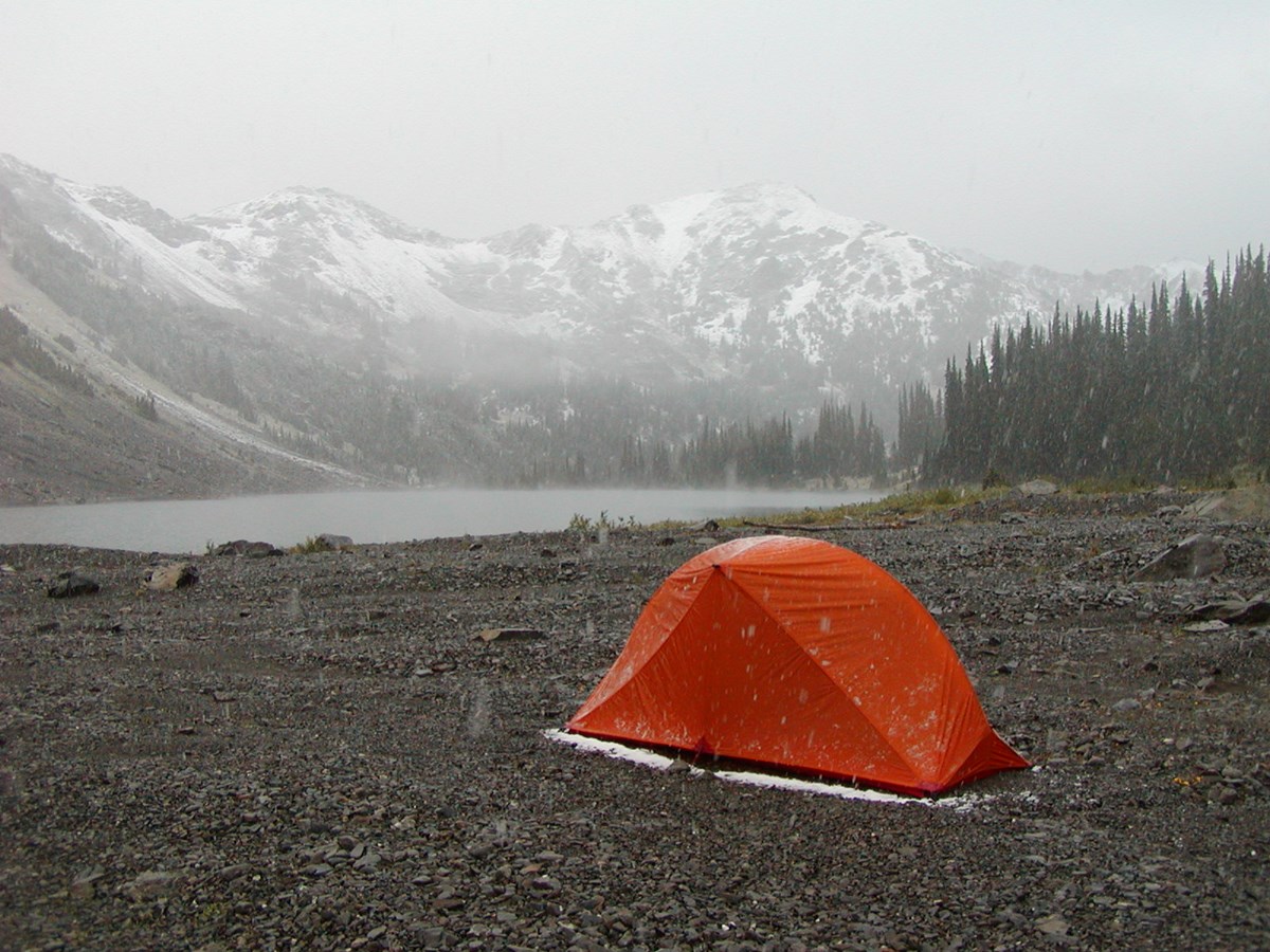 A tent in light snow on a rocky lakeshore, surrounded by snow tipped mountains