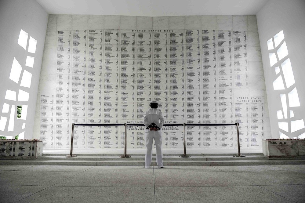 Shrine Room of the USS Arizona Memorial In the Shrine Room at the USS Arizona Memorial, a large wall lists the names of the Arizona’s dead. A person in uniform stands facing the walll, holding a folded American flag.