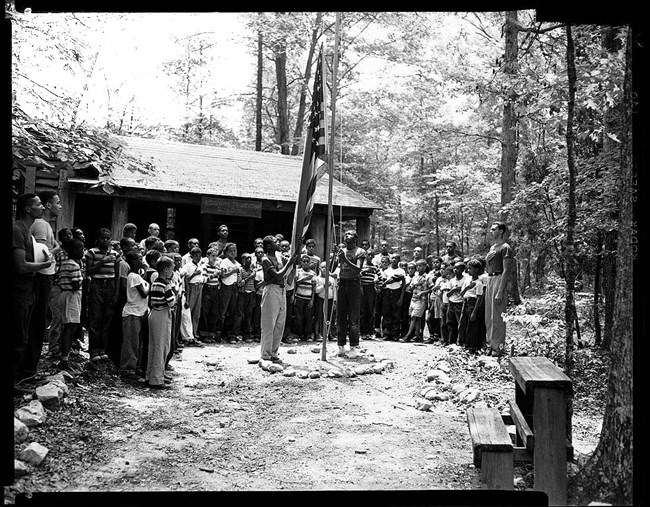 Historic photo of a semi-circle of young men at Camp Lichtman, arching around a clearing with a flagpole beside a cabin