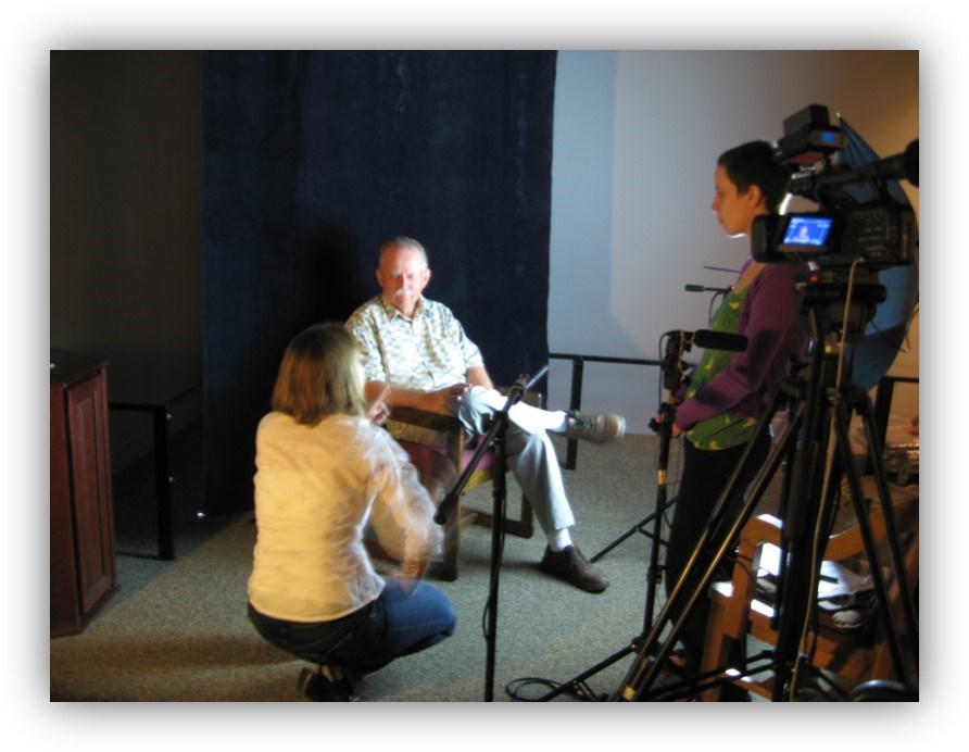 An interviewer kneels beside a man seated in a chair. Another person stands beside a camera, microphones, and recording equipment.