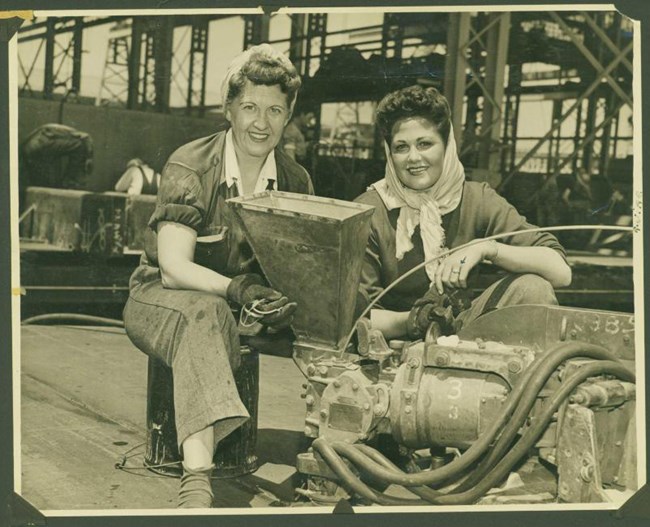 Two women in coveralls and scarves over their hair work on an engine