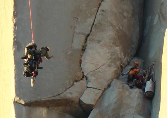 Two people hang from a rope on a cliff face, where two others stand on a ledge during a search and rescue operation.
