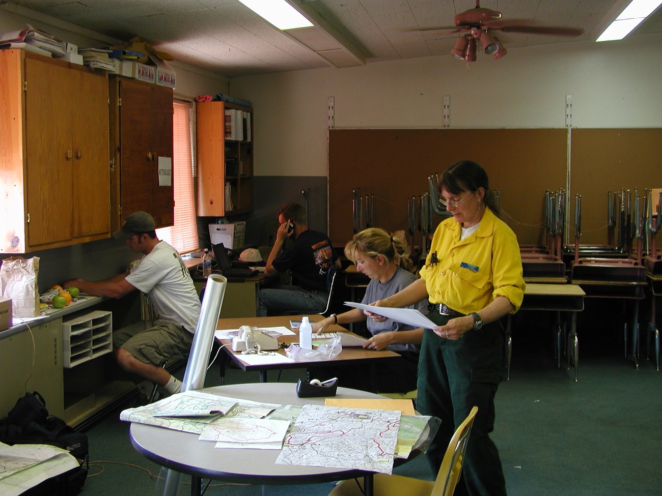 Mary Kwart, standing beside a table of maps, looks at a document in her hands. Three other people work in the background.