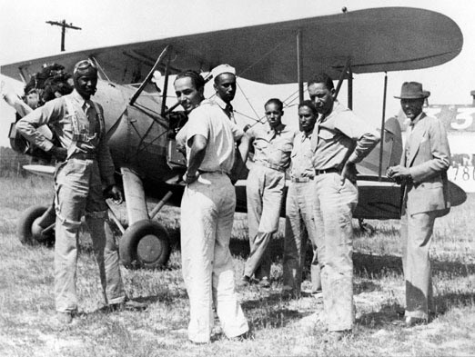 Tuskegee A group of airmen stand near the wing of a biplane in a grassy airfield.