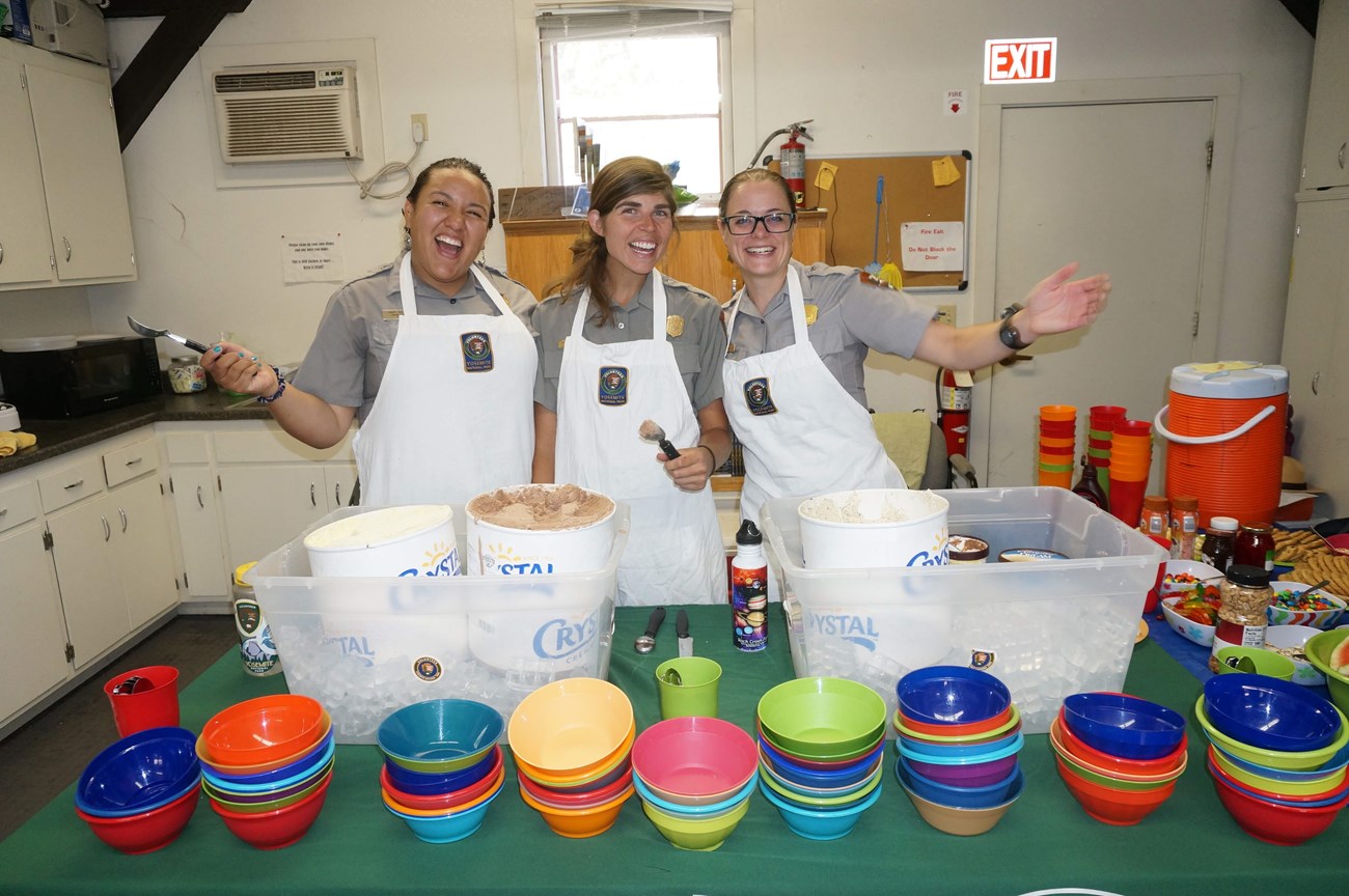 Three people in NPS uniform and aprons smile from behind a table with tubs of ice cream, stacks of colorful bowls, and ice cream toppings.