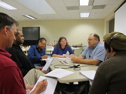 A group of people seated around a table records an interview with a man during an oral history workshop