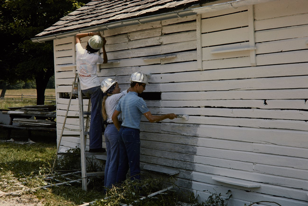 Three teenagers, one on a ladder, in the Youth Conservation Corps work on painting a building at Gettysburg National Military Park.