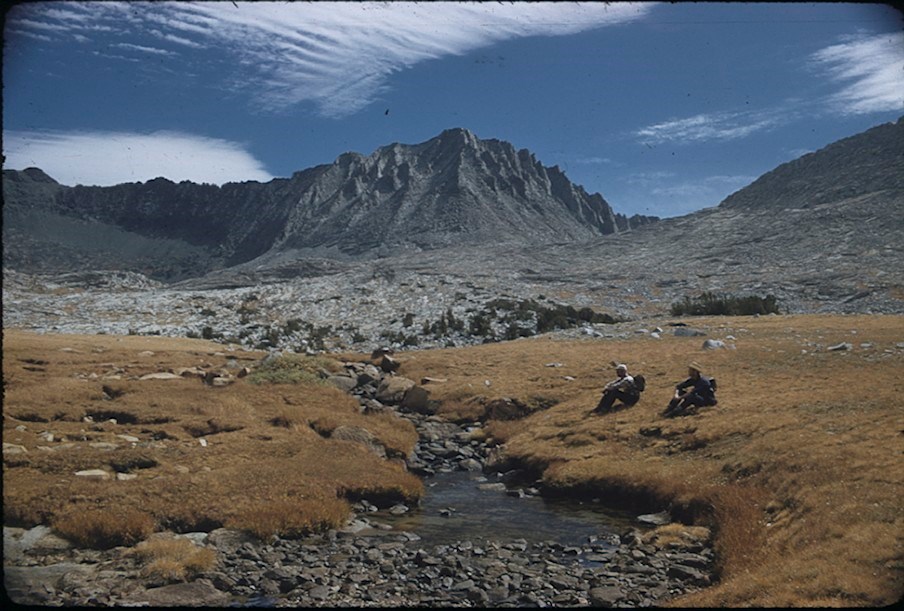 Two people sit on low grass alongside a creek, flowing from rocky mountains in the background