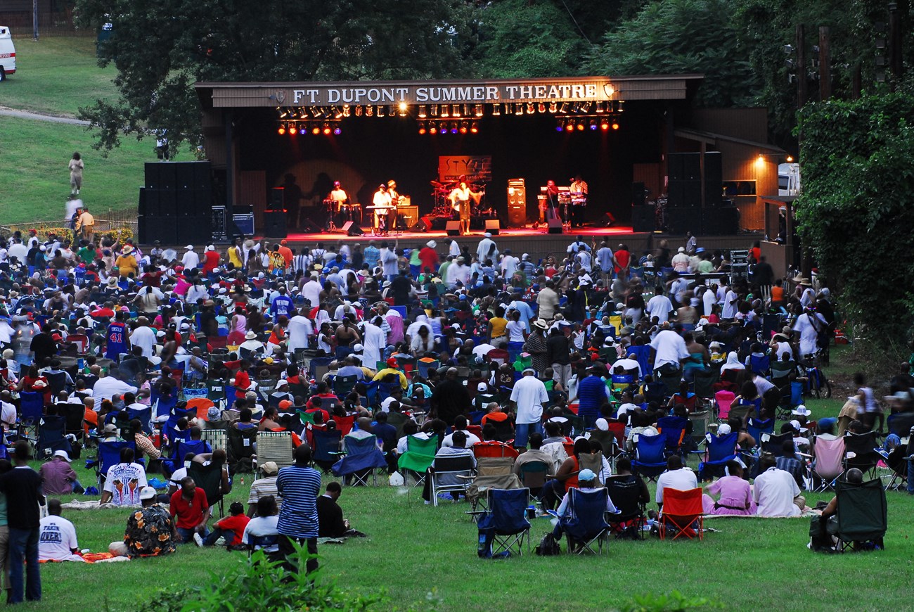 Fort Dupont Summer Theater A large crowd is gathered in the grass in front of an outdoor stage that says "Fort Dupont Summer Theater"