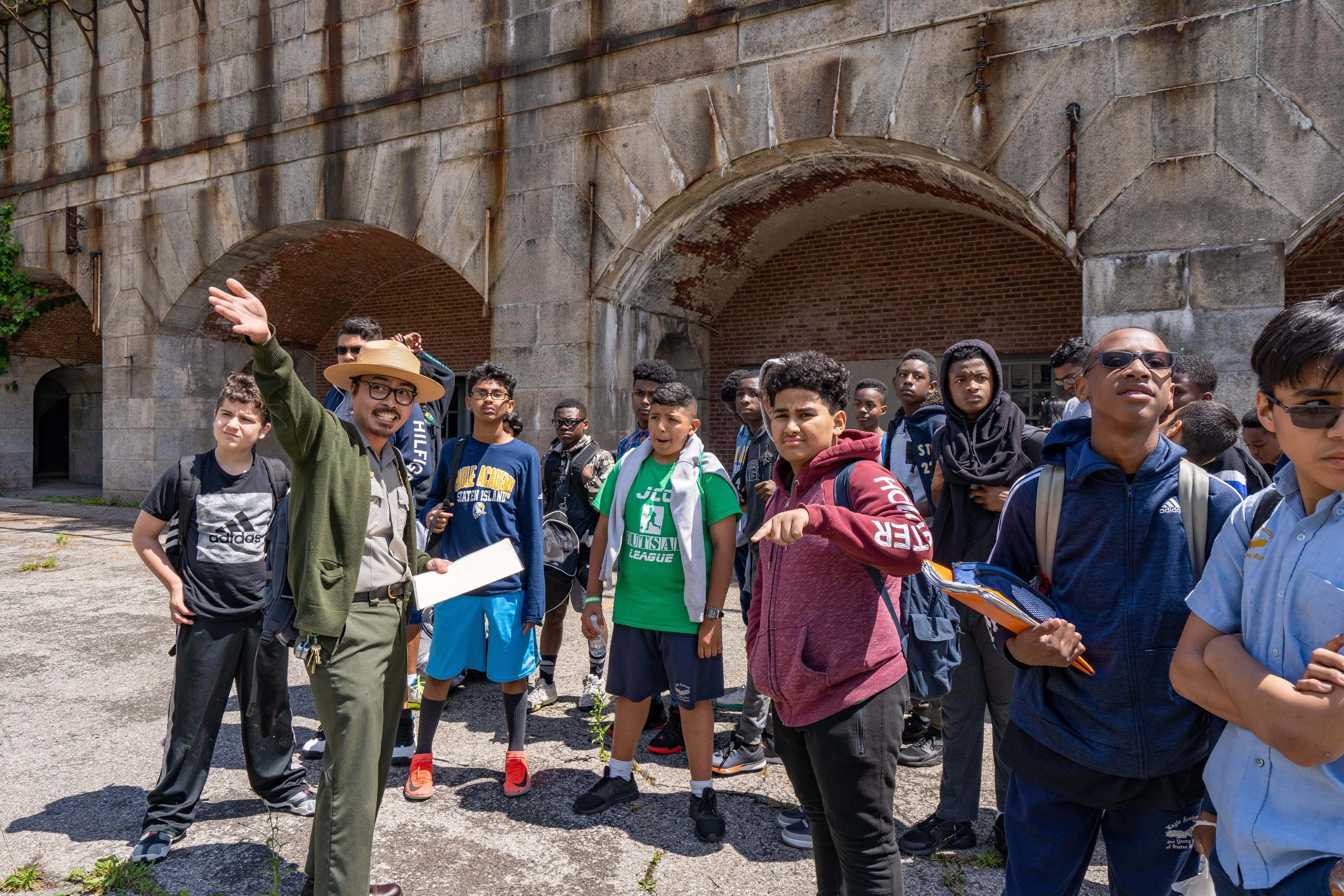 A park ranger gestures with outstretched arm, while a class of teenage boys looks on. A fort with arching openings in the background.