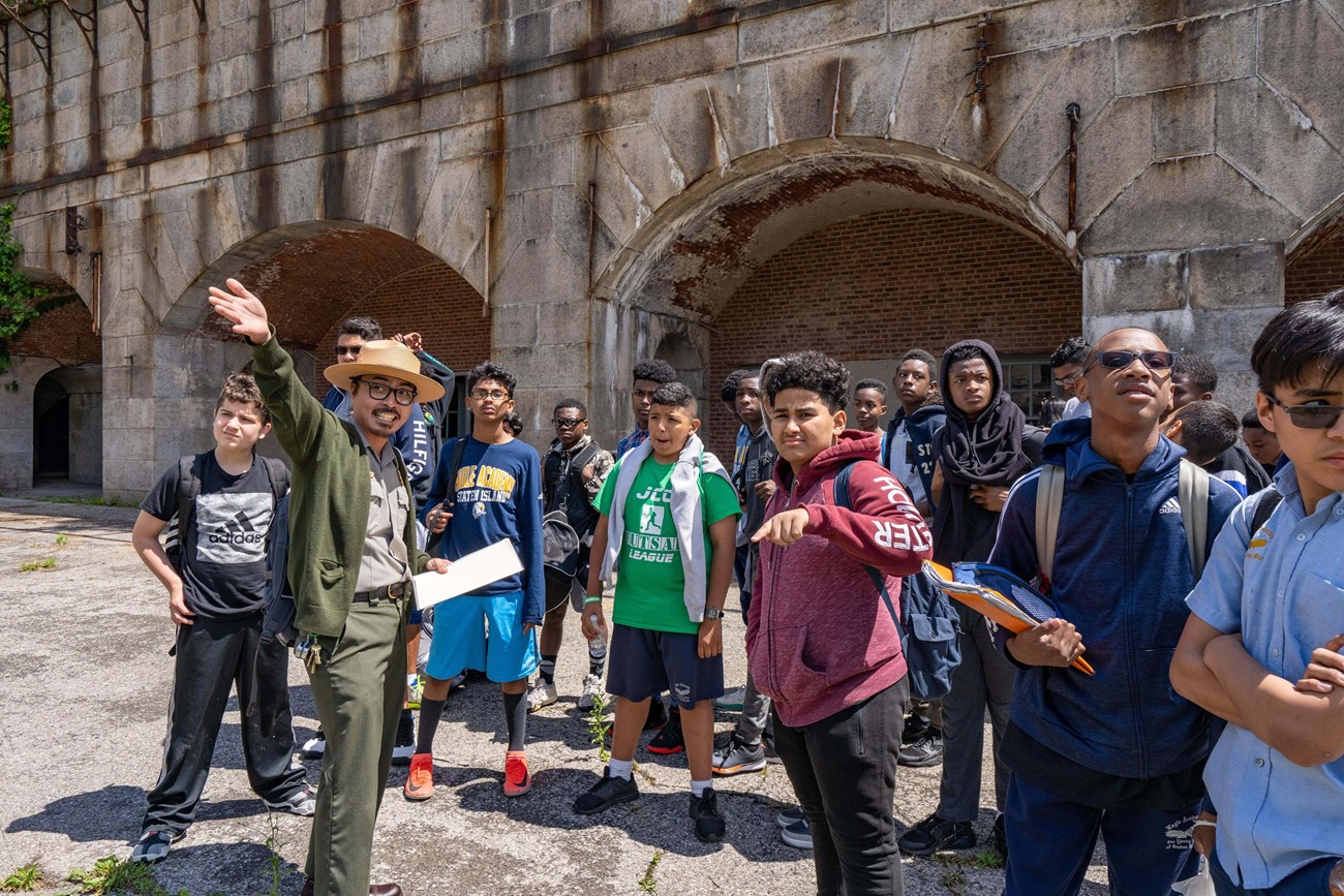 A park ranger gestures with outstretched arm, while a class of teenage boys looks on. A fort with arching openings in the background.