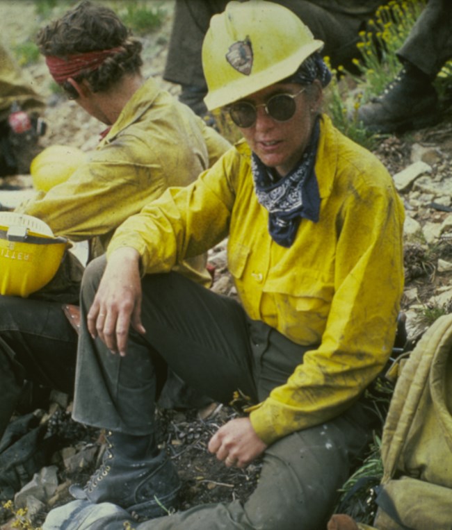 Mary Kwart sits on the ground near other crew members, with a hardhat, sunglasses, bandana around her neck, and firefighting uniform.