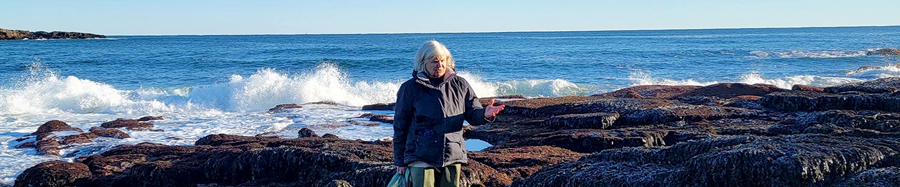Woman with white hair and a warm coat gesturing as she speaks, as waves crash on algae-covered rocks right behind her.