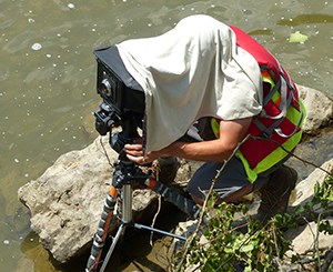 Contractor with large-format camera at Cuyahoga Valley National Park. Image credit: NPS A man in an orange and green safety vest crouches on rocks in a river while he looks through a large camera on a tripod. His head is covered with a cloth.