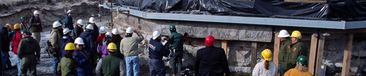 About 20 people in hard hats and winter clothing look up at the base of a building. Snow is on the ground around them.