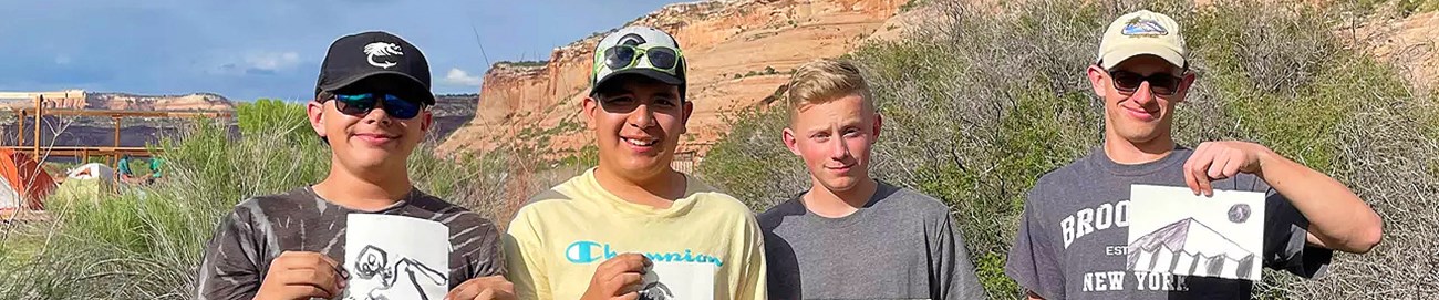 Four young men holding up black and white paintings while standing in front of red-rock canyons and green trees.