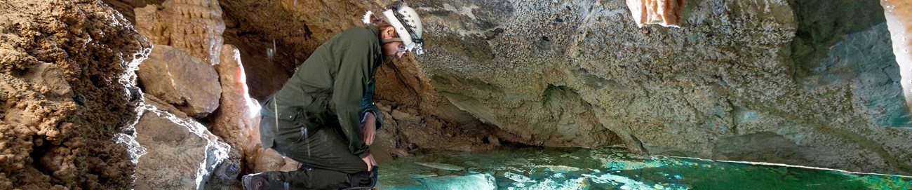 A man in an NPS uniform with a helmet and headlamp looks over a turquoise pool surrounded by cave formations