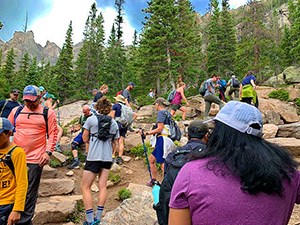 A group of people hiking up a steep, boulder-covered slope with evergreens and blue skies in the background