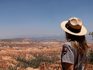 A woman with long hair and in an NPS uniform and hat stands with her back to the camera as she looks at a distant landscape filled with red rocks