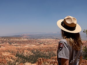 Looking at the hoodoos in Bryce Canyon A woman with long hair and in an NPS uniform and hat stands with her back to the camera as she looks at a distant landscape filled with red rocks