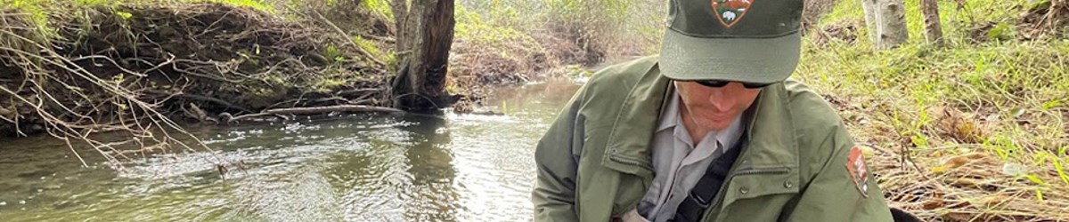 A man wearing an NPS uniform and sunglasses looks down next to a creek.