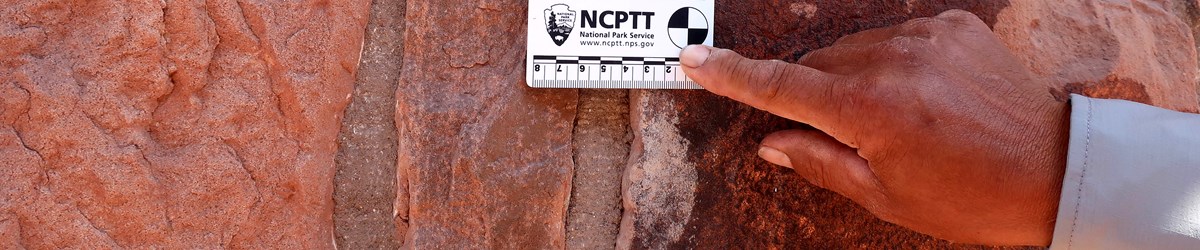 Mortar sampling at Hubble Trading Post. A man's hand holds a scale card against a mortar joint in an old building.