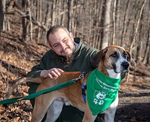 Ranger with BARK ranger at Catoctin Mountain Park. Image credit: NPS / Catoctin Mountain Park A light brown and white dog on a green leash, wearing a green BARK ranger bandana, stands in front of a crouching park ranger with a beard. A winter forest with leafless trees is behind them.
