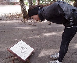Reader at Mount Rainier National Park. Image credit: NPS A woman wearing sneakers, black tights, gray jacket and black headband bends over to read a sign almost buried in mud. Behind her are trees, a river and a bridge.