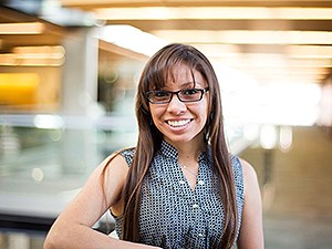 A young woman with long, brown hair and glasses, wearing a blue checkered top, miles at the camera.