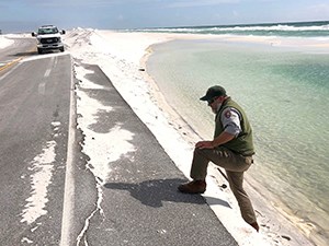 Road damaged from Hurricane Sally in 2020. Man in NPS uniform climbs onto road from sandy beach.