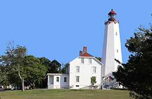 Sandy Hook Lighthouse, Gateway National Recreation Area. Image credit: NPS White lighthouse with a red bell tower next to a white house with a red roof on a green lawn next to green trees with a blue sky