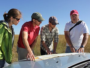 Three men and a woman look at a chart resting on the hood of a car. The older man on the right is explaining something to the others. Behind them are a large grassy field and blue skies.