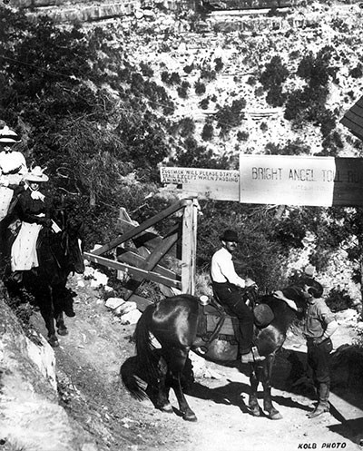 Riders pay a toll at Cameron's Bright Angel Trailhead around 1910. Gate with toll sign: "Footman will please stand on trail except when passing animals." Corner of Kolb studio building on right.