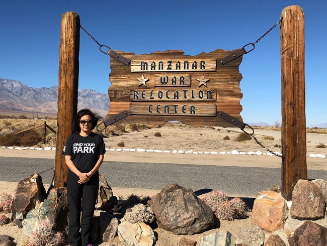 Under a deep blue sky and with mountains in the background a woman stands next to a sign that says Manzanar War Relocation Center.