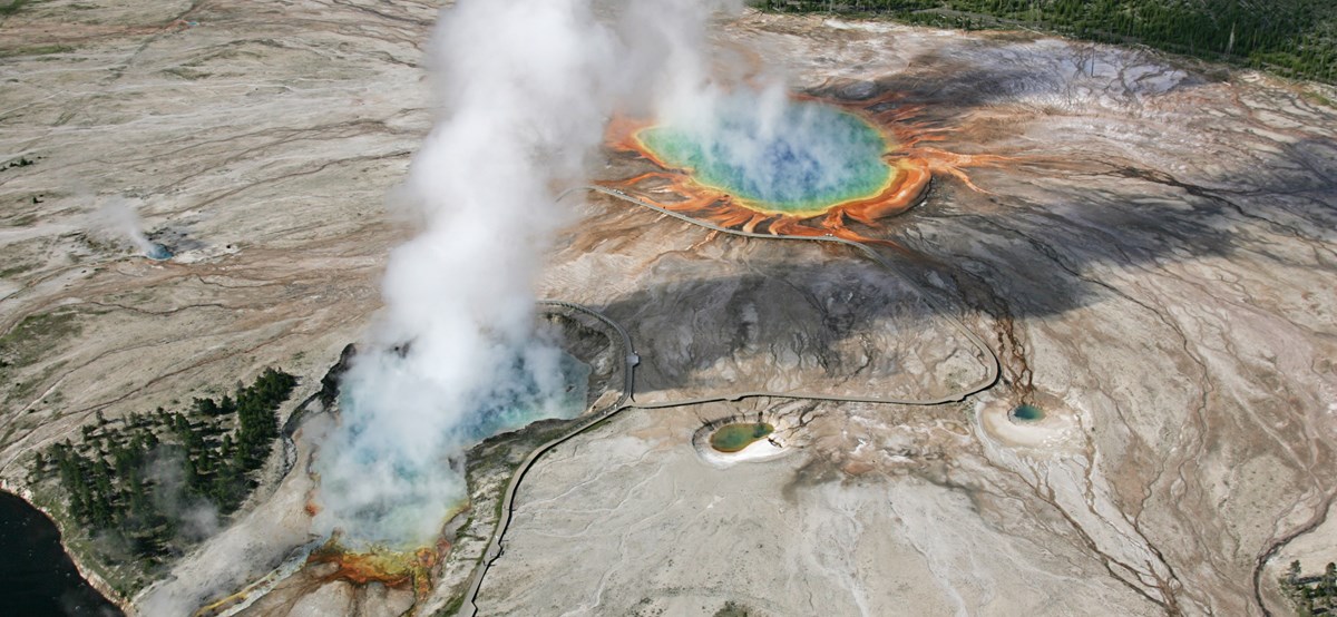 An aerial view of two rainbow colored hot springs giving off steam.  
