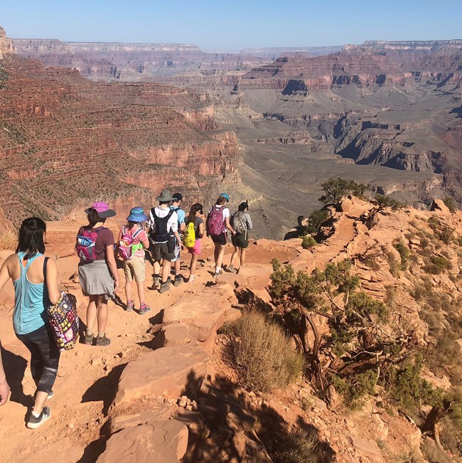 a group of people walking single-file down an unpaved trail switchback in the side of a cliff. In the distance, colorful peaks rising out of a deep canyon.