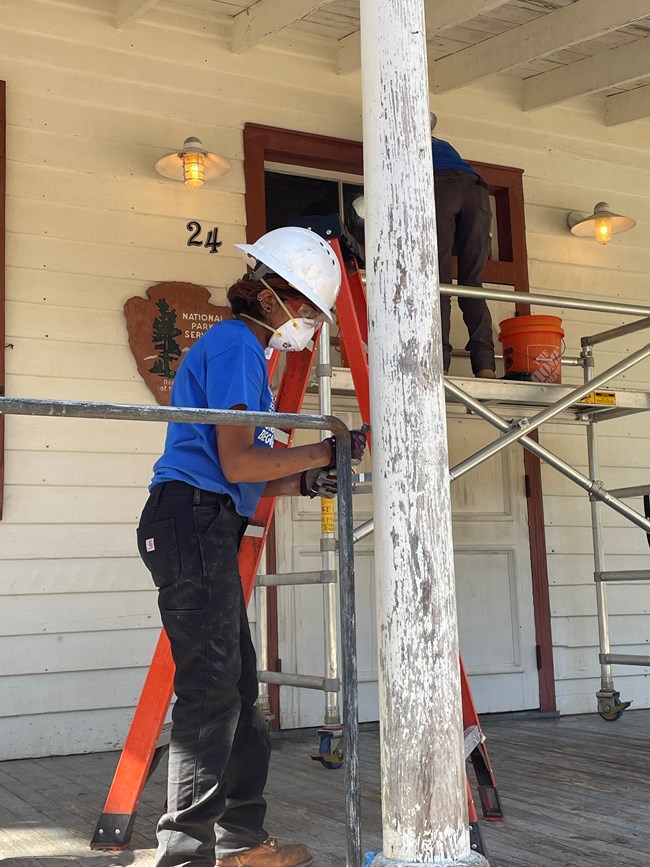 SCA Women's Team A woman wearing personal protection equipment works on scraping paint off a column on the porch of Darrah Hall.