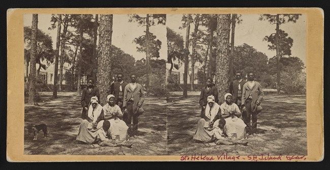 A Family on St. Helena Island During the Reconstruction era Three men stand, two women, and a child sit outside in front of pine trees and houses.