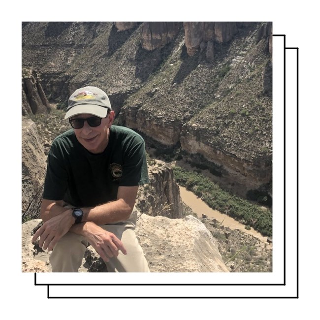 A smiling older man in ball cap and sunglasses kneels on the rim of a canyon