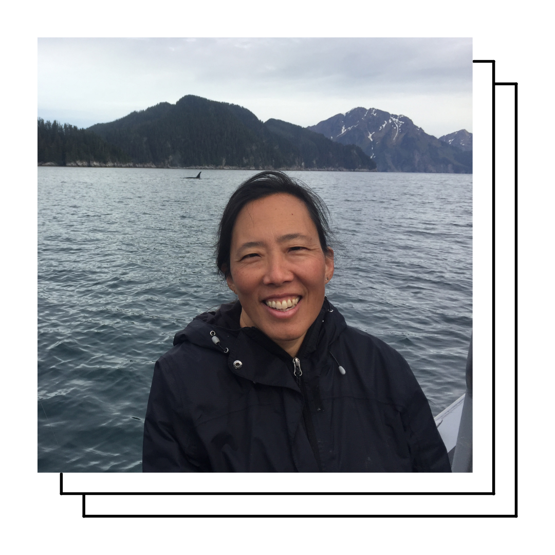 A smiling woman with dark hair and tan skin with the ocean, an orca fin, and mountains in the background