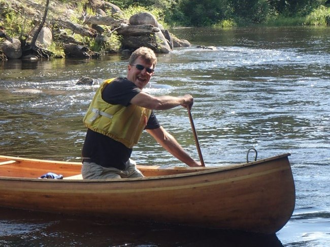 a smiling man wearing a life jacket paddles a canoe on a river