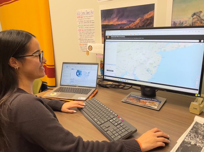 A young woman smiles and stands at her desk in front of a computer with the 2024 NRI data viewer on the screen.