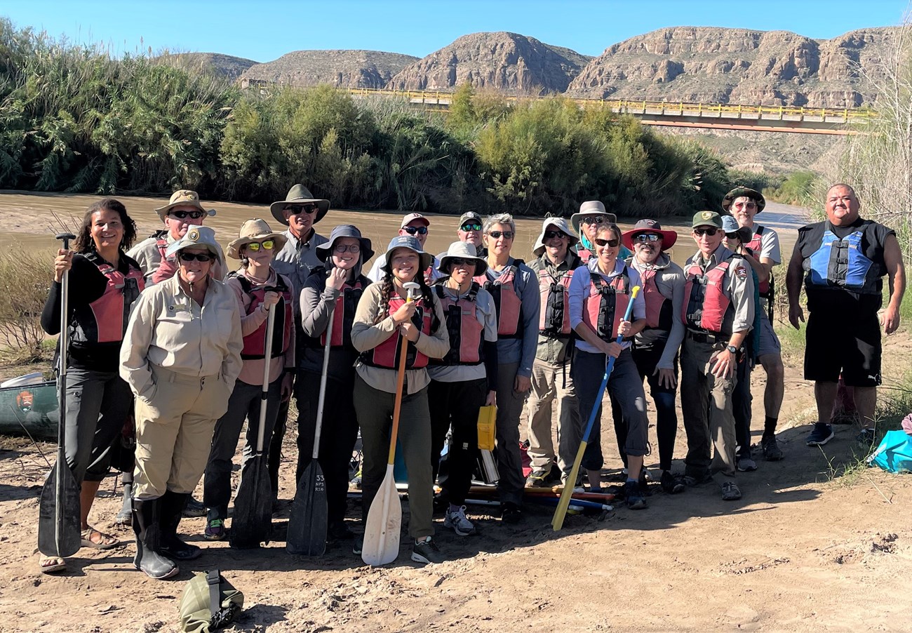 A group of people wearing lifejackets and holding oars stand next to a river