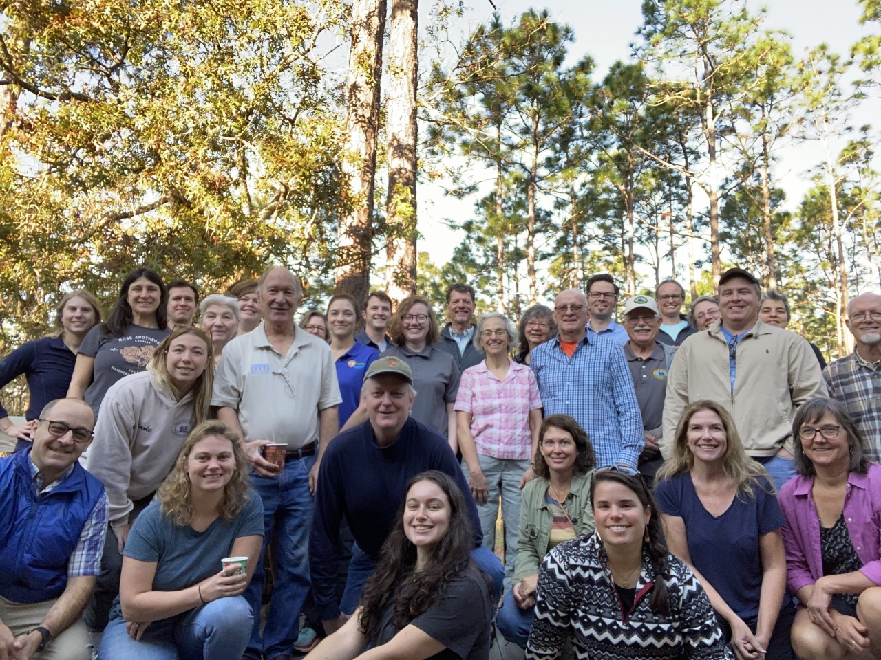 a group of around 30 smiling people of varying ages pose for a photo in the forest