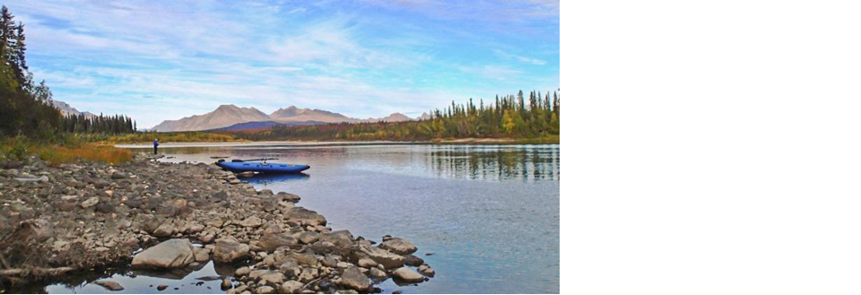 Lone kayaker standing on river bank looking at mountains.