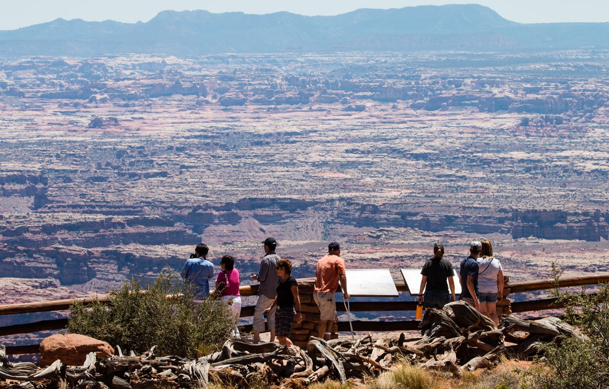 Several visitors look out over an expanse of canyons in the desert. The visitors are standing behind an informational sign and fence.
